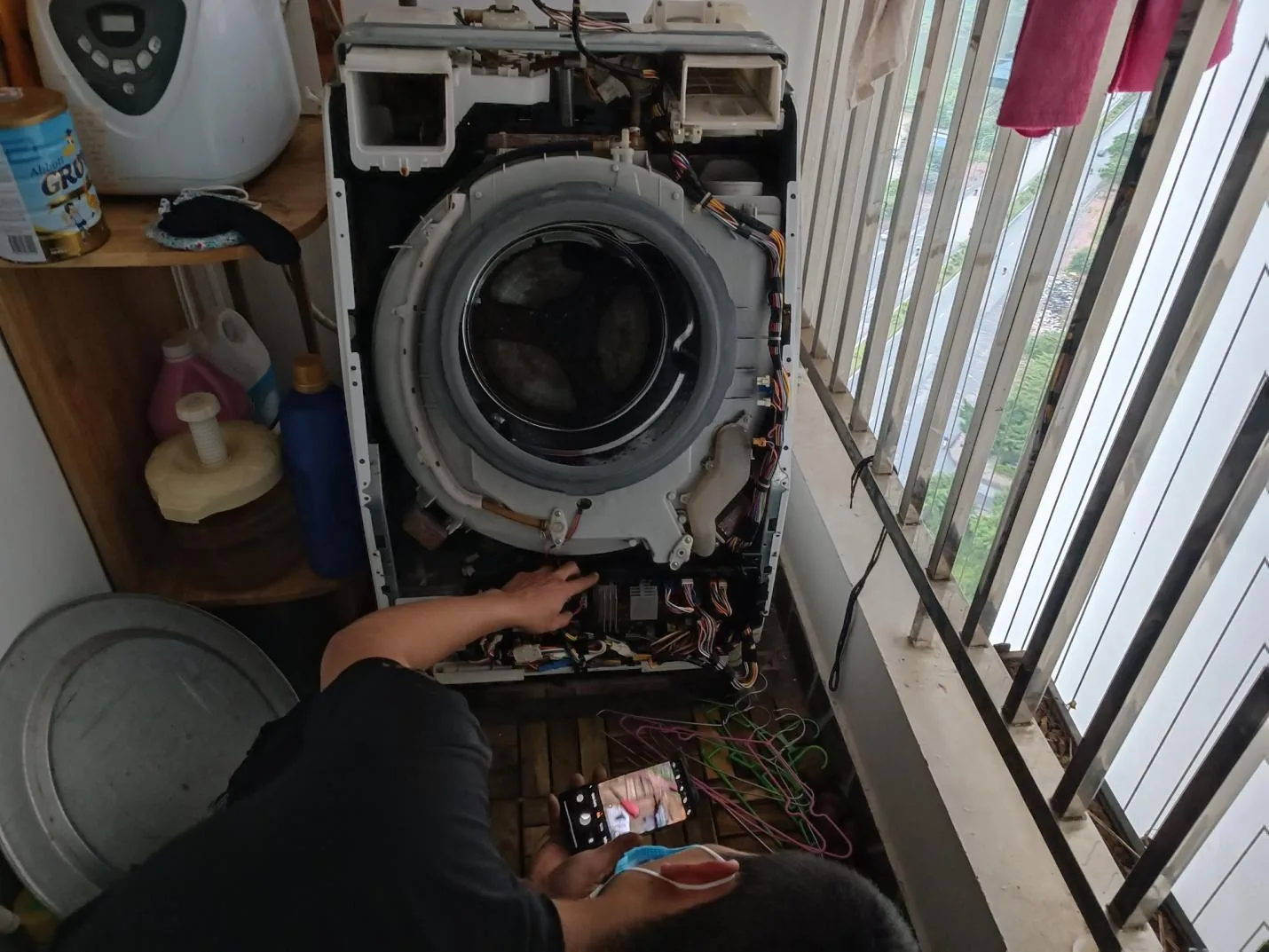 Thợ tháo mặt trước máy giặt cửa ngang tại ban công chung cư, lộ thùng ngoài, gioăng cửa và bó dây điện