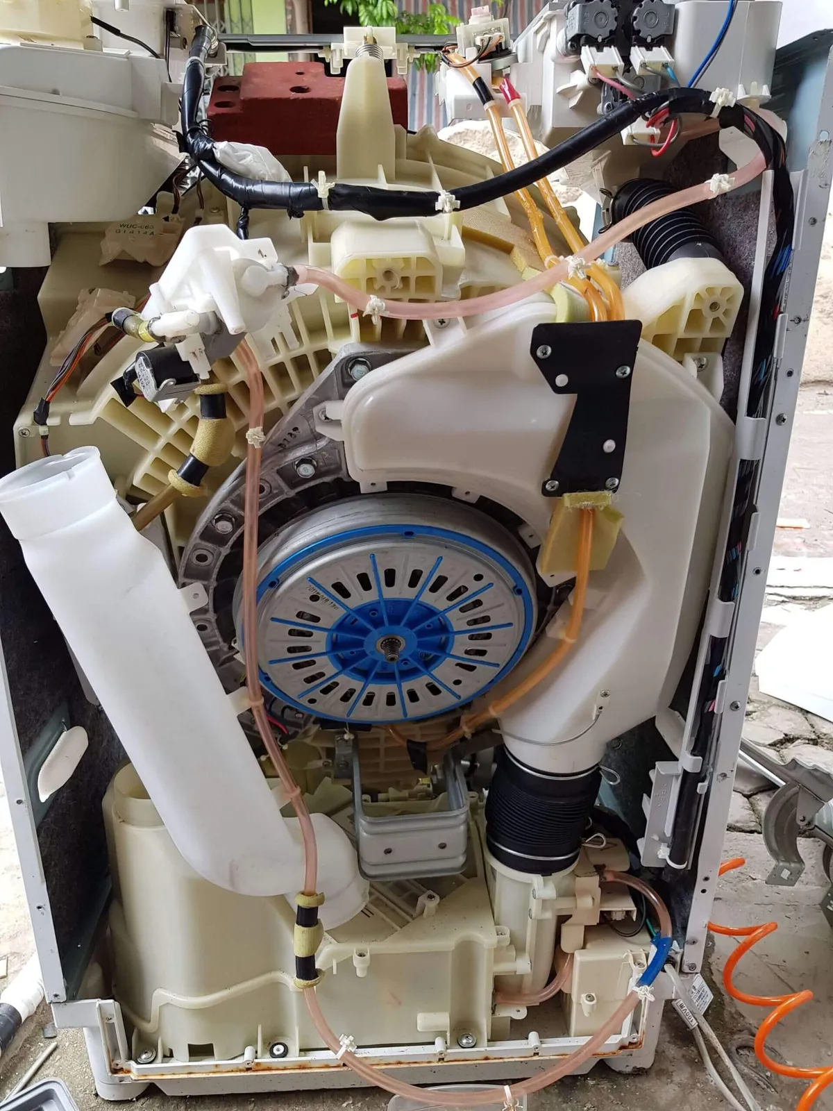 Mặt sau máy giặt lồng đứng tháo vỏ, lộ rotor truyền động trực tiếp, thùng ngoài và ống cảm biến mực nước